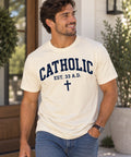 Model wearing a natural Catholic Varsity T-shirt with navy lettering, EST. 33 A.D. text, and a cross.