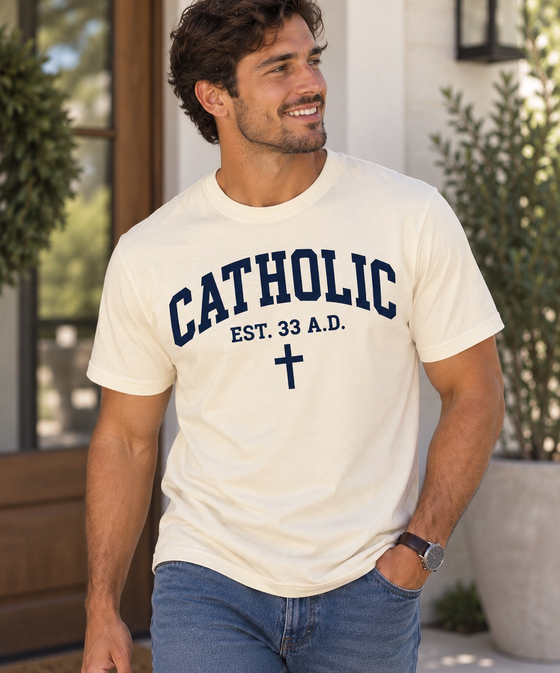 Model wearing a natural Catholic Varsity T-shirt with navy lettering, EST. 33 A.D. text, and a cross.
