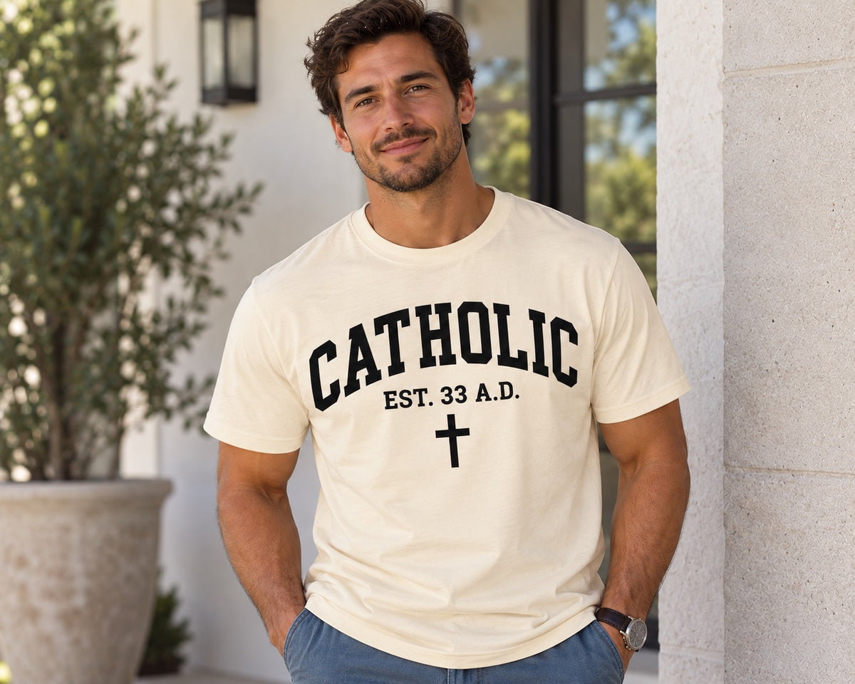 Model wearing a natural Catholic Varsity T-shirt with black lettering, EST. 33 A.D. text, and a cross.