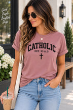 Model wearing a pink T-shirt with black Catholic lettering, EST. 33 A.D. text, and a cross.