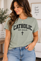 Model wearing a pale green T-shirt with black Catholic lettering, EST. 33 A.D. text, and a cross.