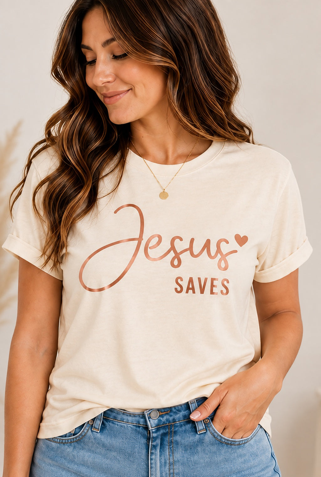 Model wearing a natural-colored Jesus Saves T-shirt with pink/copper-colored script lettering and a heart.