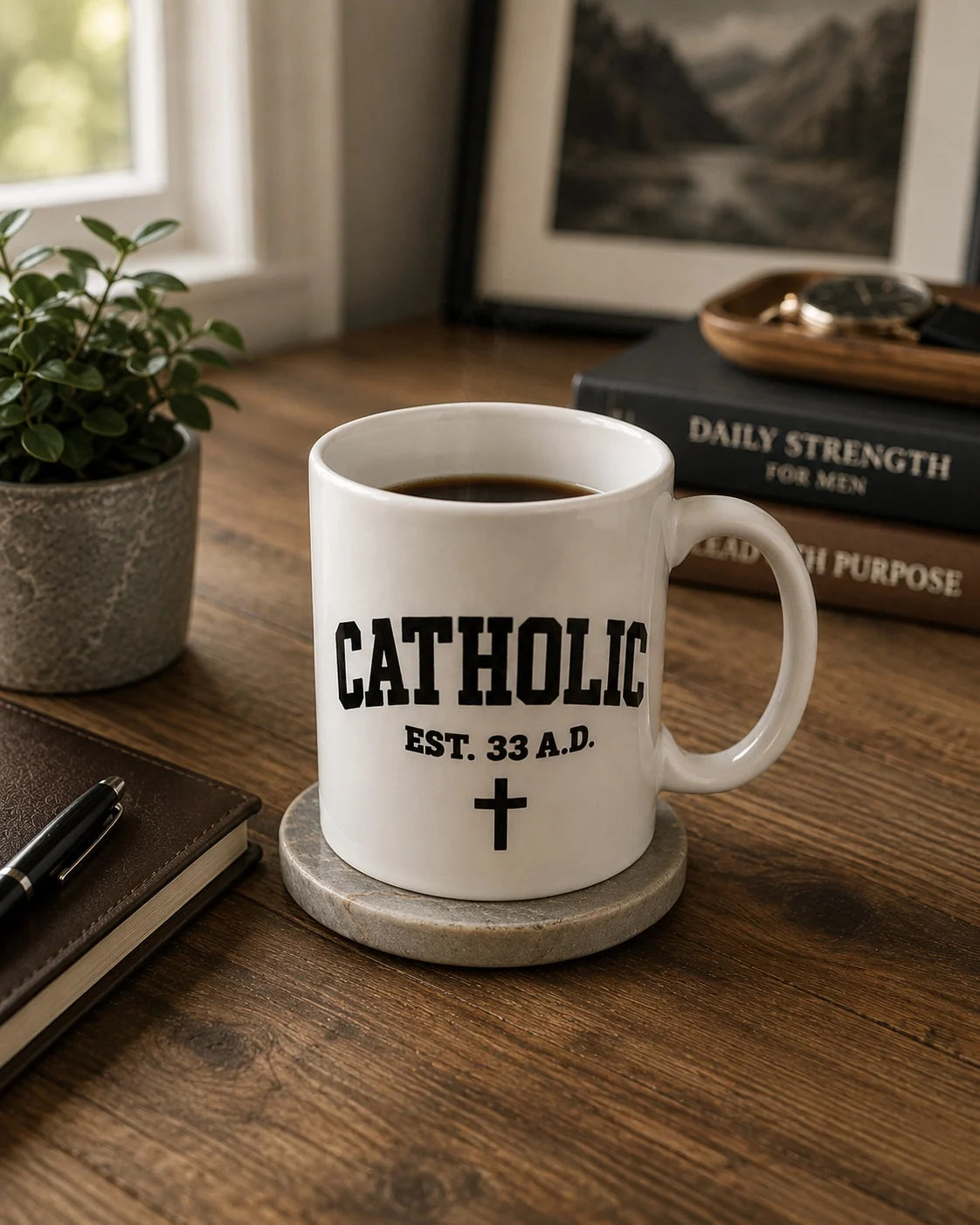 White Catholic Varsity Mug on a wooden table beside books, a notebook, and greenery